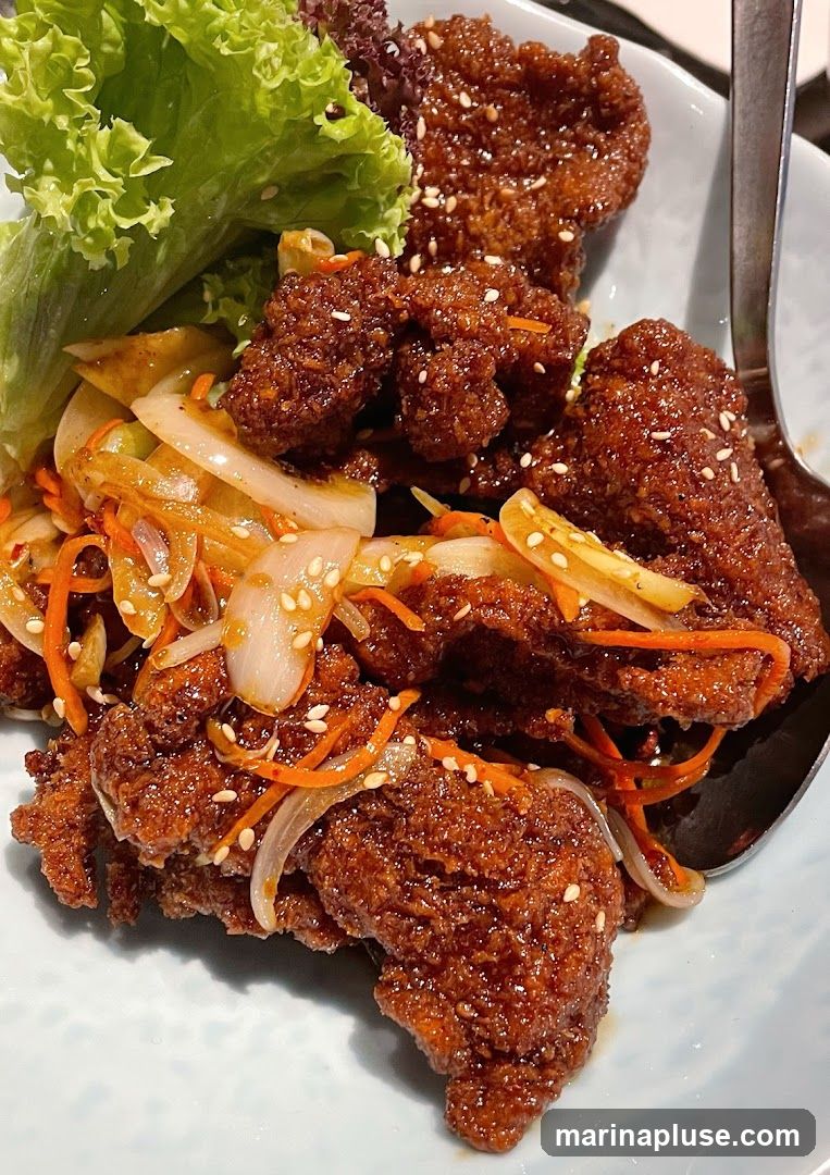 A close-up shot of a dish featuring crispy, glazed fried chicken pieces served with thinly sliced onions, carrots, and lettuce. The chicken is coated in a dark, glossy sauce and sprinkled with sesame seeds.