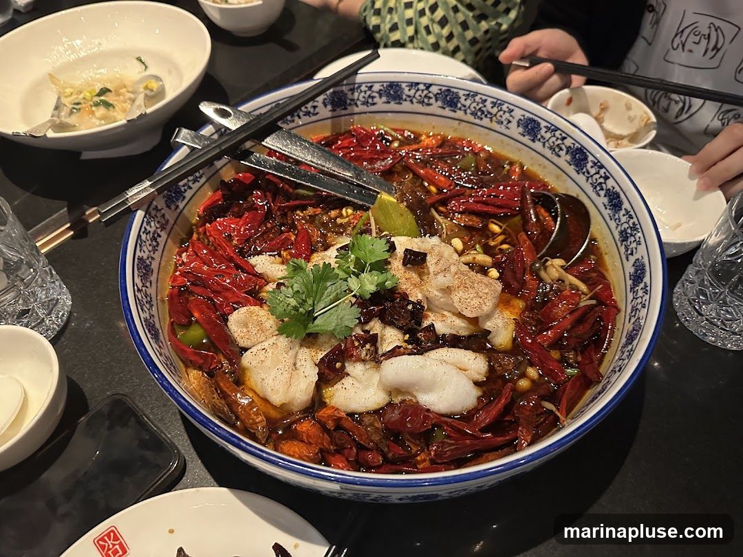 A close-up overhead shot of a large bowl of fish in a spicy broth, filled with dried red chilies, Sichuan peppercorns, and garnished with cilantro. Chopsticks are resting on the rim of the bowl. Several small white bowls and glasses are visible around the main dish, suggesting a shared meal.
