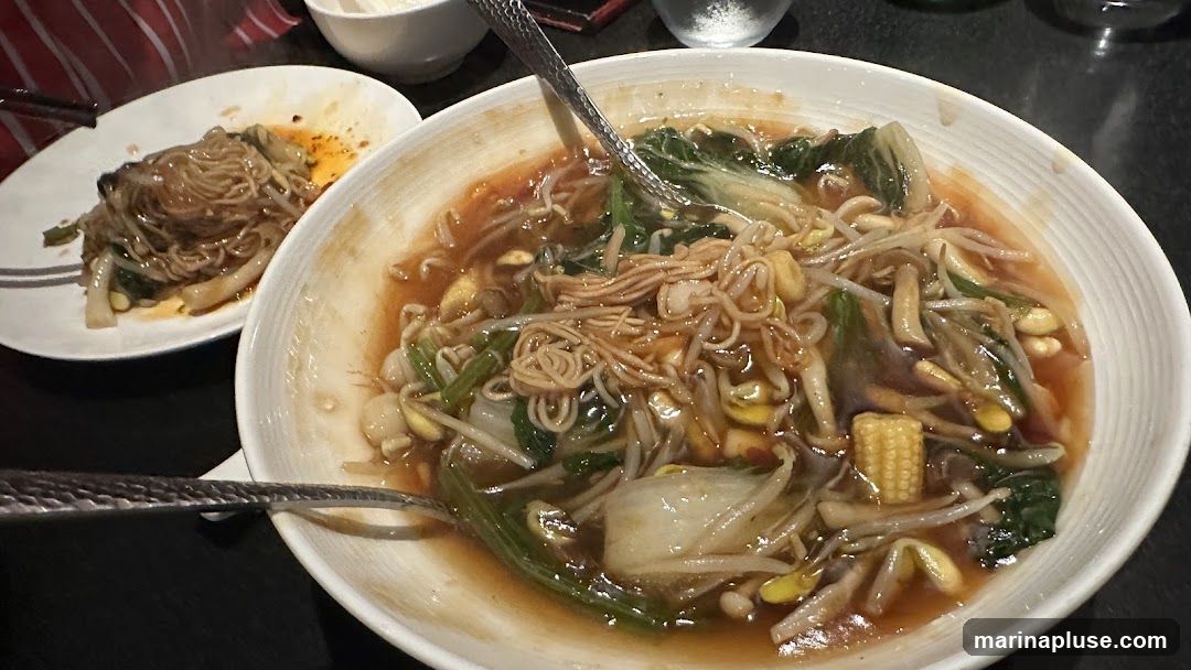 A close-up shot of two bowls of food on a dark table. The bowl in the foreground is larger and contains a noodle dish with broth, bok choy, bean sprouts, mushrooms, and baby corn. A spoon is in the bowl. To the left, a smaller plate holds a portion of noodles with vegetables.