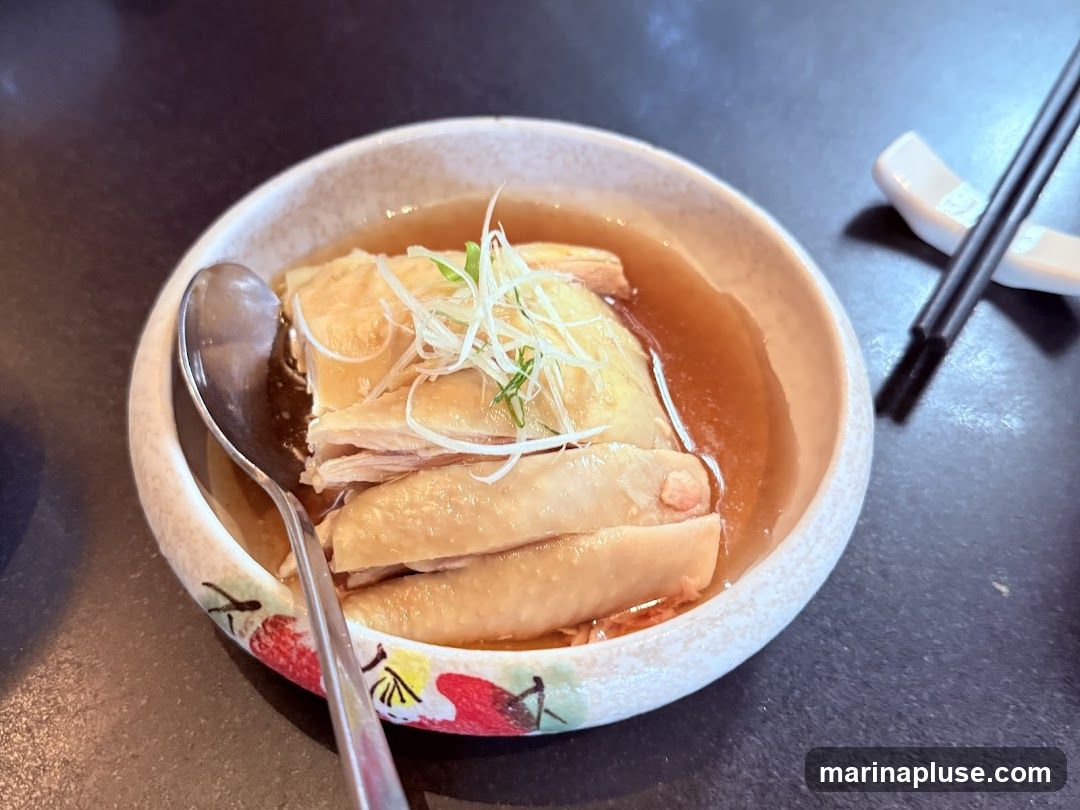 A close-up shot of a bowl of poached chicken, garnished with shredded scallions. The chicken is served in a light broth. A spoon rests in the bowl, and chopsticks are visible in a holder to the right. The bowl has a decorative rim with floral patterns.