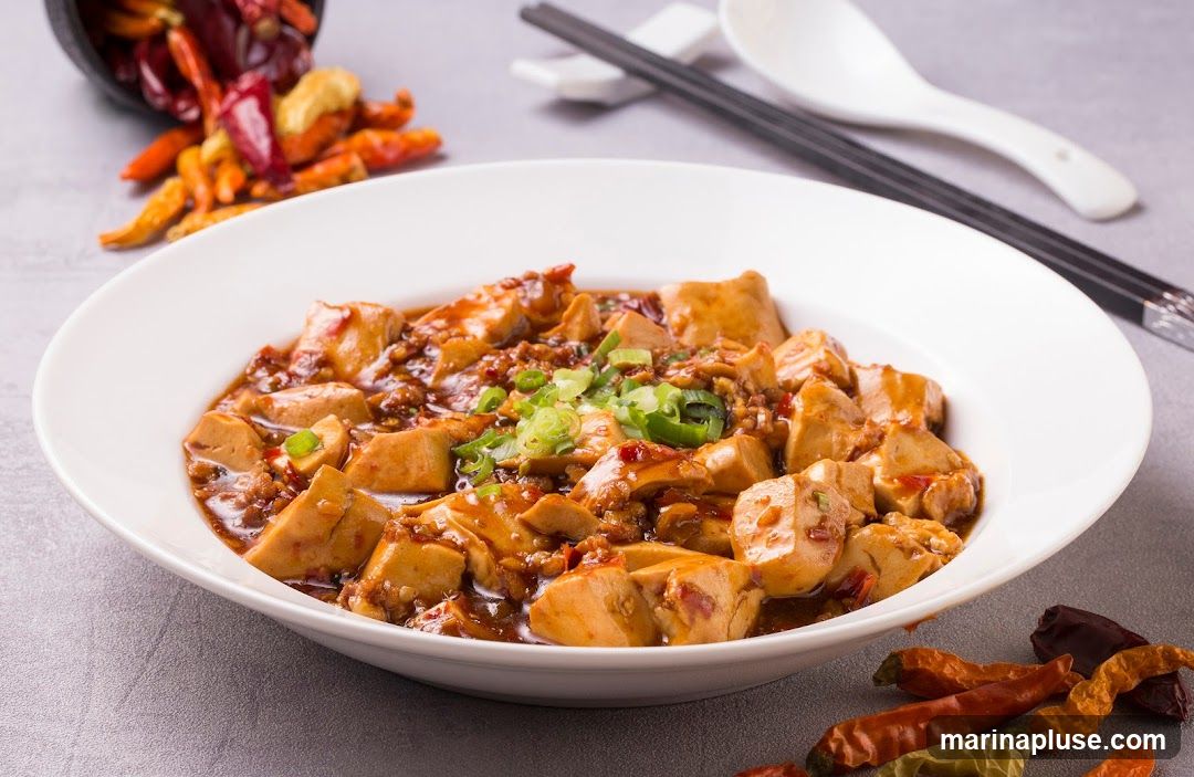 A close-up shot of Mapo Tofu in a white bowl, garnished with chopped green onions. Dried chili peppers are scattered around the bowl and in a small black container in the background. Chopsticks and a spoon rest on white chopstick holders behind the bowl.