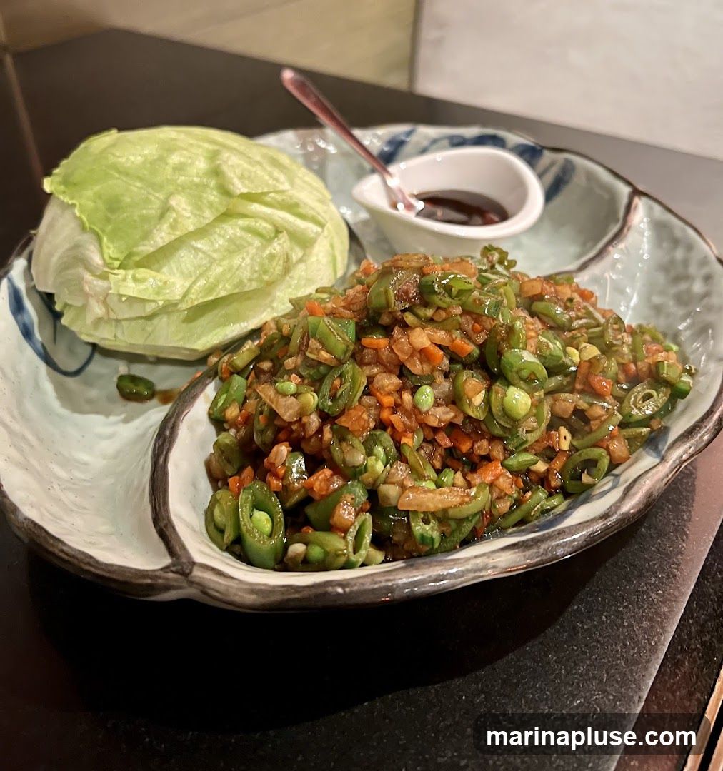 A close-up shot of a dish featuring a mound of chopped green beans, carrots, and other vegetables, served with a small dish of dark sauce and a head of lettuce. The dish is presented on a decorative plate with blue and white patterns.