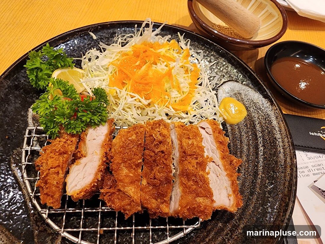 A plate of sliced, breaded and fried pork cutlet is served with shredded cabbage, grated carrots, a lemon wedge, parsley, a dollop of mustard, and a side of dipping sauce. A small bowl with a wooden pestle and seeds, and another small bowl with sauce are also visible. A menu is partially visible on the right.