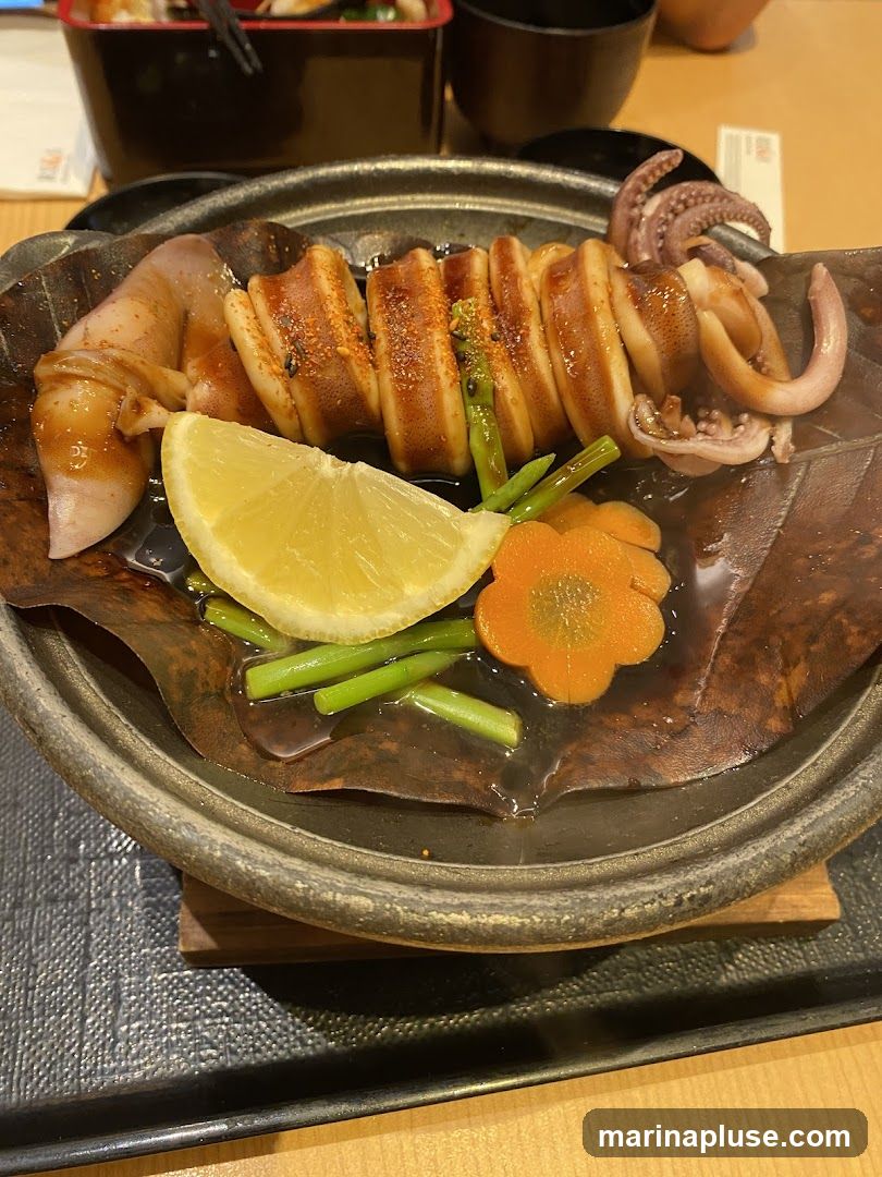 A close-up shot of a dish featuring grilled squid, sliced and arranged on a dark, textured platter. The squid is seasoned with spices and served with a lemon wedge, green beans, and a carrot slice shaped like a flower. The dish appears to be in a restaurant setting, with other food items and bowls visible in the blurred background.