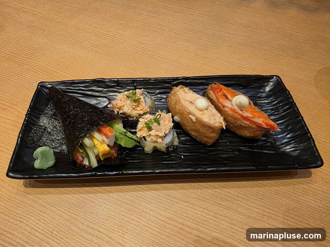 A black rectangular plate holds a selection of sushi and inari. On the left is a hand roll (temaki) filled with crab, cucumber, egg, and avocado. Next to it are two pieces of sushi topped with a creamy crab mixture and green onions. To the right are two pieces of inari sushi, one filled with a creamy mixture and topped with a white dollop, and the other filled with imitation crab sticks and a white dollop. A small dollop of green wasabi is on the side. The food is arranged on a wooden table.