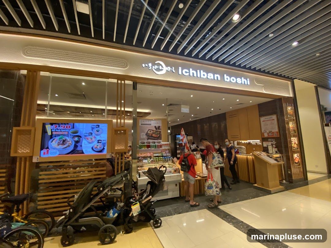 The exterior of the Ichiban Boshi restaurant features a prominent sign with the restaurant's name in English and Japanese characters. A large digital screen displays food items and a special offer of $7.90 Off Peak Specials. The entrance is wide, with glass windows allowing a view into the interior. Several people are seen entering and exiting the establishment. Strollers and bicycles are parked outside the entrance.