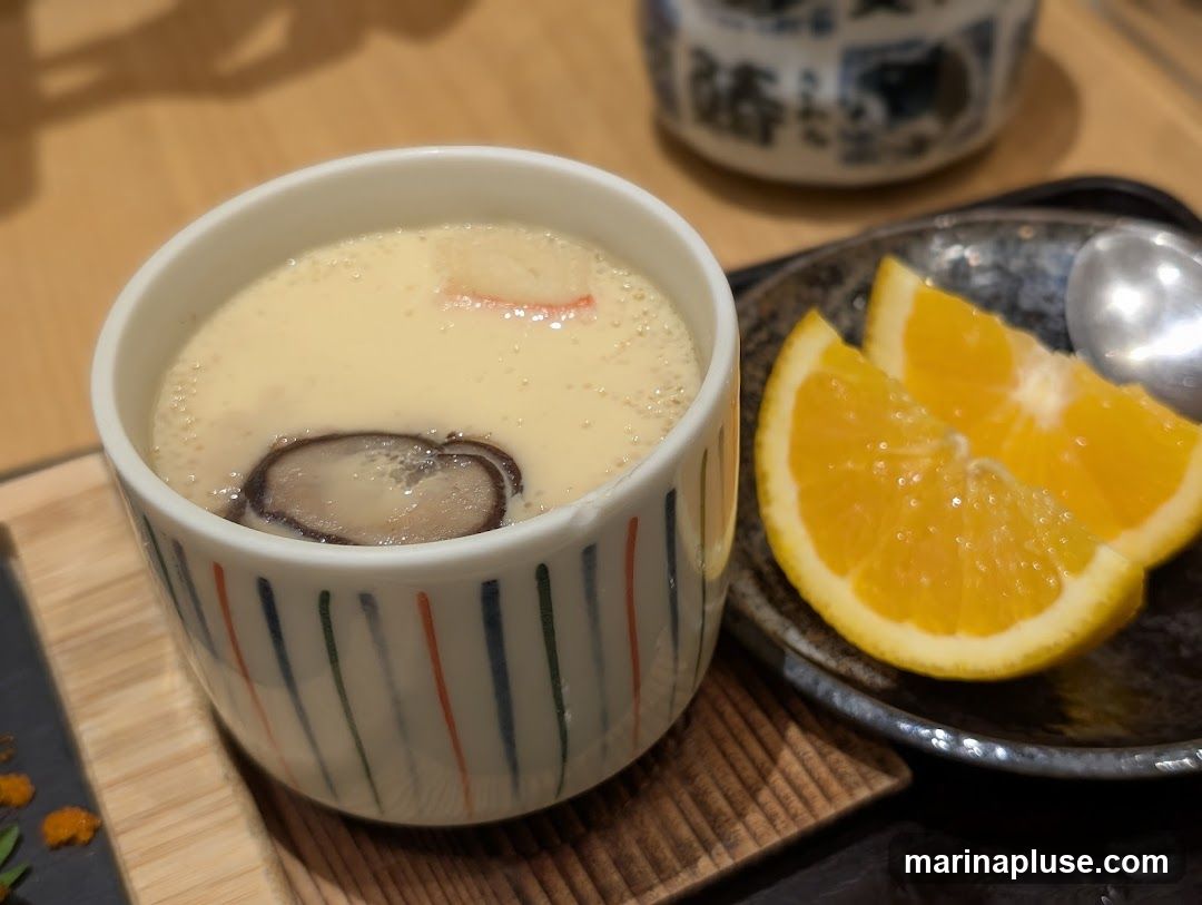A close-up shot of a Japanese savory egg custard, known as Chawanmushi, served in a decorative ceramic cup. The custard is smooth and creamy, topped with a slice of shiitake mushroom and a piece of red imitation crab stick. To the right, two bright orange slices are presented on a dark, textured plate alongside a silver spoon. The dish is placed on a wooden surface, with a blurred background suggesting a restaurant setting.