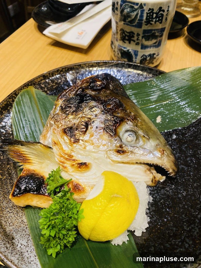 A close-up shot of a grilled salmon head served on a dark, textured plate. The fish is charred in places, with visible grill marks. It is garnished with a lemon wedge and a sprig of parsley, resting on a bed of shredded daikon radish and a large green leaf. In the background, chopsticks and a decorative cup with Japanese characters are visible on a wooden table.