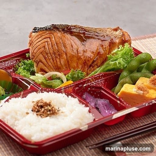 A bento box filled with a grilled salmon fillet, rice, edamame, tamagoyaki (rolled omelet), and pickled vegetables. The bento box is red with a decorative pattern.