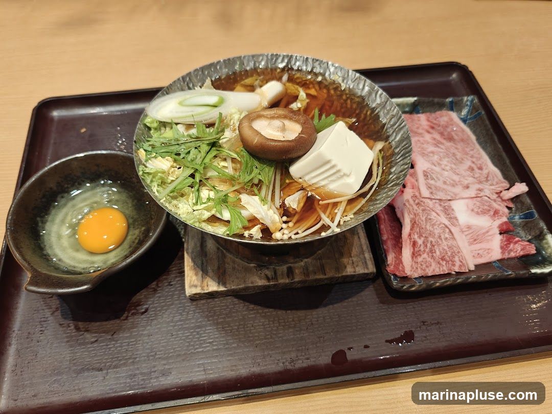 A close-up shot of a sukiyaki dish served on a dark tray. The dish features thinly sliced beef, tofu, napa cabbage, mushrooms, and scallions in a savory broth. A small bowl with a raw egg yolk is placed to the left of the main dish.