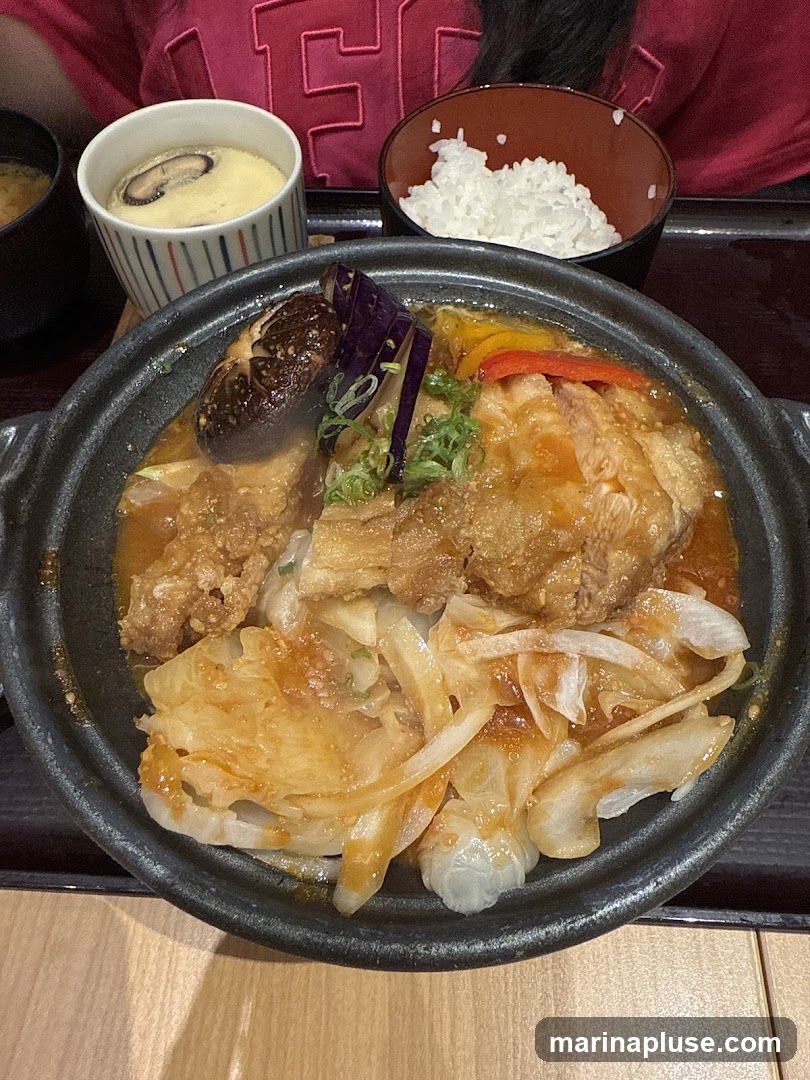 A close-up overhead view of a Japanese meal. The main dish is a hot pot with meat, cabbage, onions, mushrooms, and peppers in a savory sauce. It is served with a bowl of white rice, a small bowl of miso soup, and a cup of chawanmushi (steamed egg custard) topped with a mushroom slice. The meal is presented on a dark tray. A person wearing a pink shirt with a large white 'E' graphic is visible in the background.