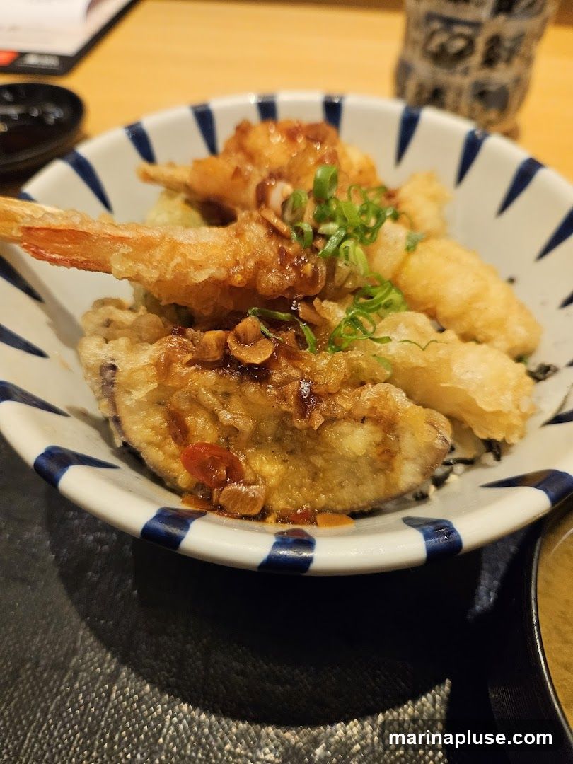 A close-up shot of a bowl filled with tempura, including shrimp and eggplant, drizzled with a savory sauce and garnished with chopped green onions. A small dish of soy sauce and a cup of miso soup are partially visible in the background.