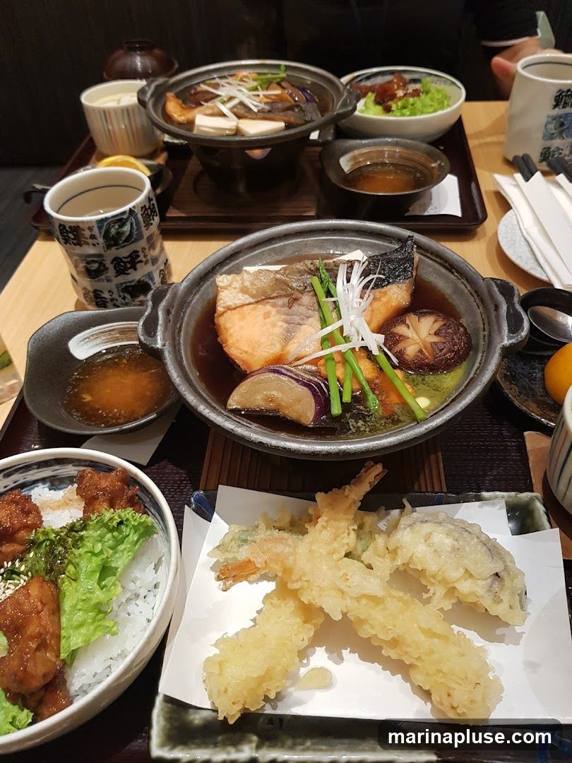 A close-up overhead view of a table laden with various Japanese dishes. In the foreground, a bowl of white rice is topped with pieces of fried chicken and lettuce. Next to it, a plate holds several pieces of tempura, including shrimp and vegetables. Further back, a simmering pot contains fish, tofu, and vegetables in a broth. Another dish features a piece of grilled fish with vegetables. A small bowl of dipping sauce and a cup of tea are also visible.