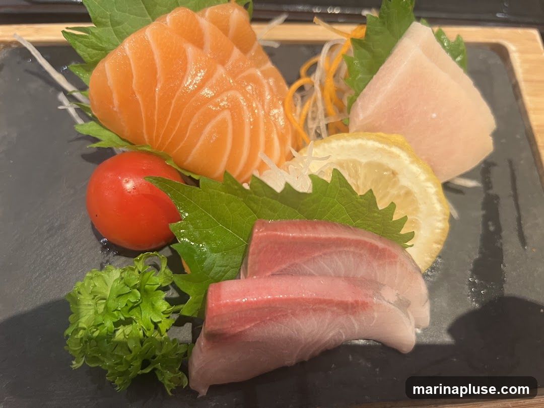 A close-up shot of a sashimi platter featuring slices of salmon, tuna, and another type of fish, garnished with a cherry tomato, lemon slice, shredded daikon radish, and parsley.