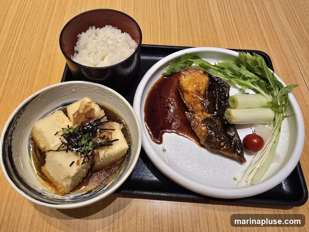 A Japanese meal set featuring grilled fish with a dark glaze, served with leeks, a cherry tomato, and a side of greens. Accompanying the fish is a bowl of white rice and a separate bowl of fried tofu cubes in broth, garnished with seaweed and green onions. The meal is presented on a black tray on a wooden table.