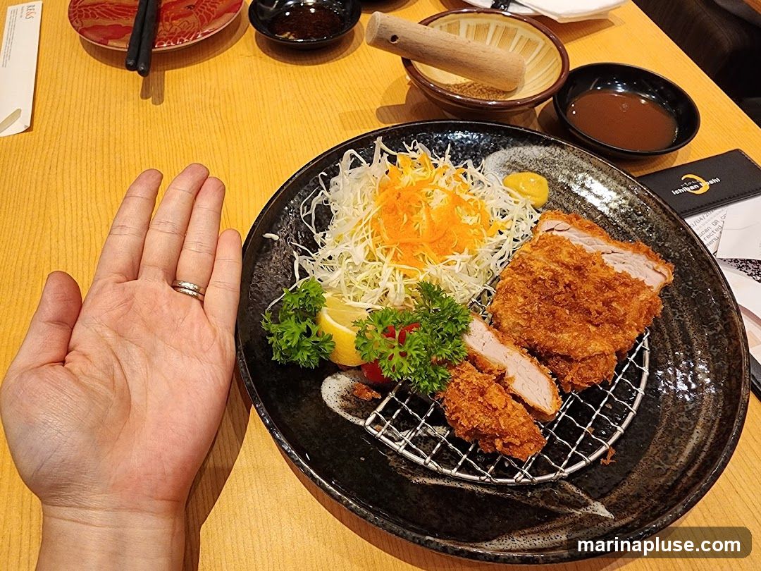 A close-up shot of a hand next to a plate of tonkatsu, which is a deep-fried pork cutlet. The tonkatsu is served with shredded cabbage, shredded carrots, a lemon wedge, and parsley. A small dish of sauce is also visible. A menu or bill is partially visible on the right side of the plate.