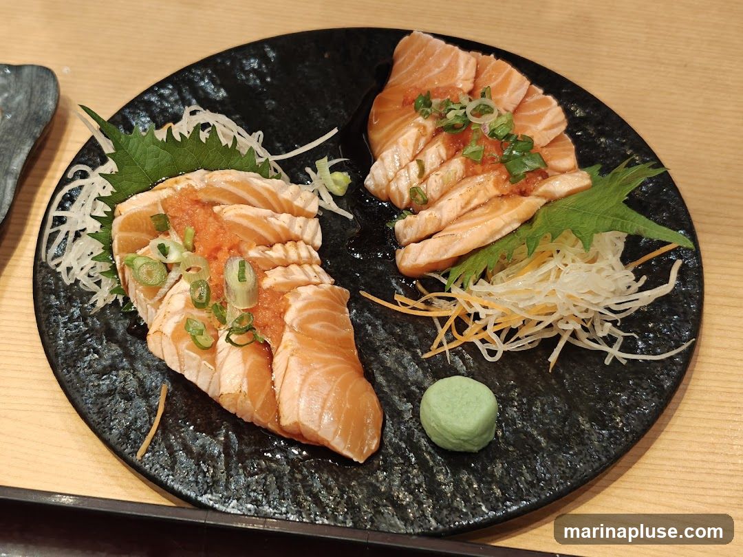A black plate holds two servings of salmon sashimi, garnished with chopped green onions and a spicy sauce. The sashimi is arranged over shredded daikon radish and julienned carrots, with a small dollop of wasabi on the side.