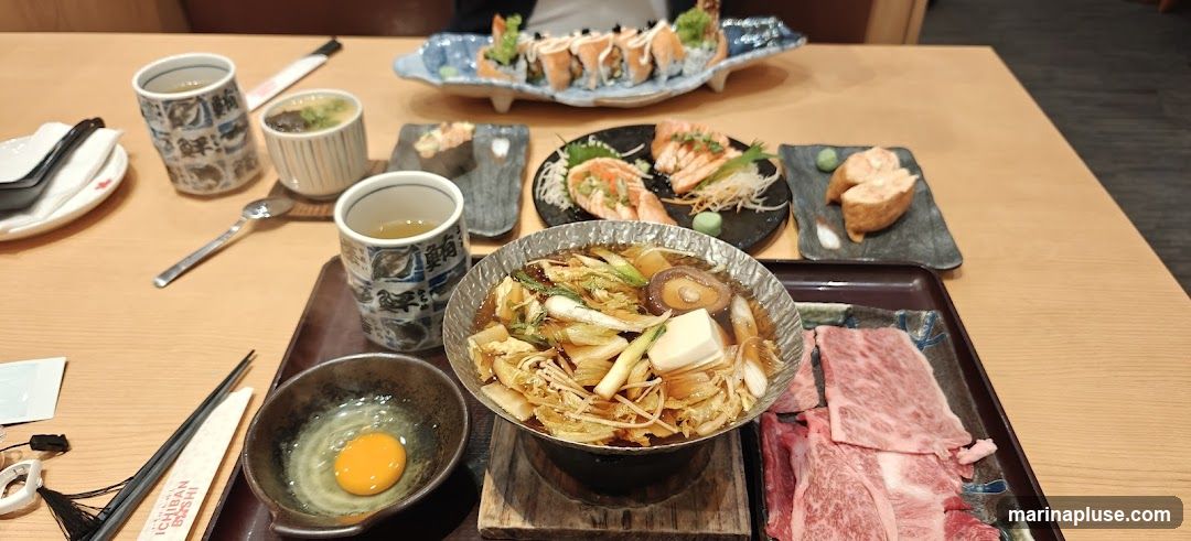 A table laden with various Japanese dishes, including a sukiyaki pot with vegetables and tofu, thinly sliced raw beef, sushi rolls, salmon sashimi, and small cups of tea or soup. A raw egg in a bowl is also visible, likely for dipping. Chopsticks and spoons are arranged on the table.