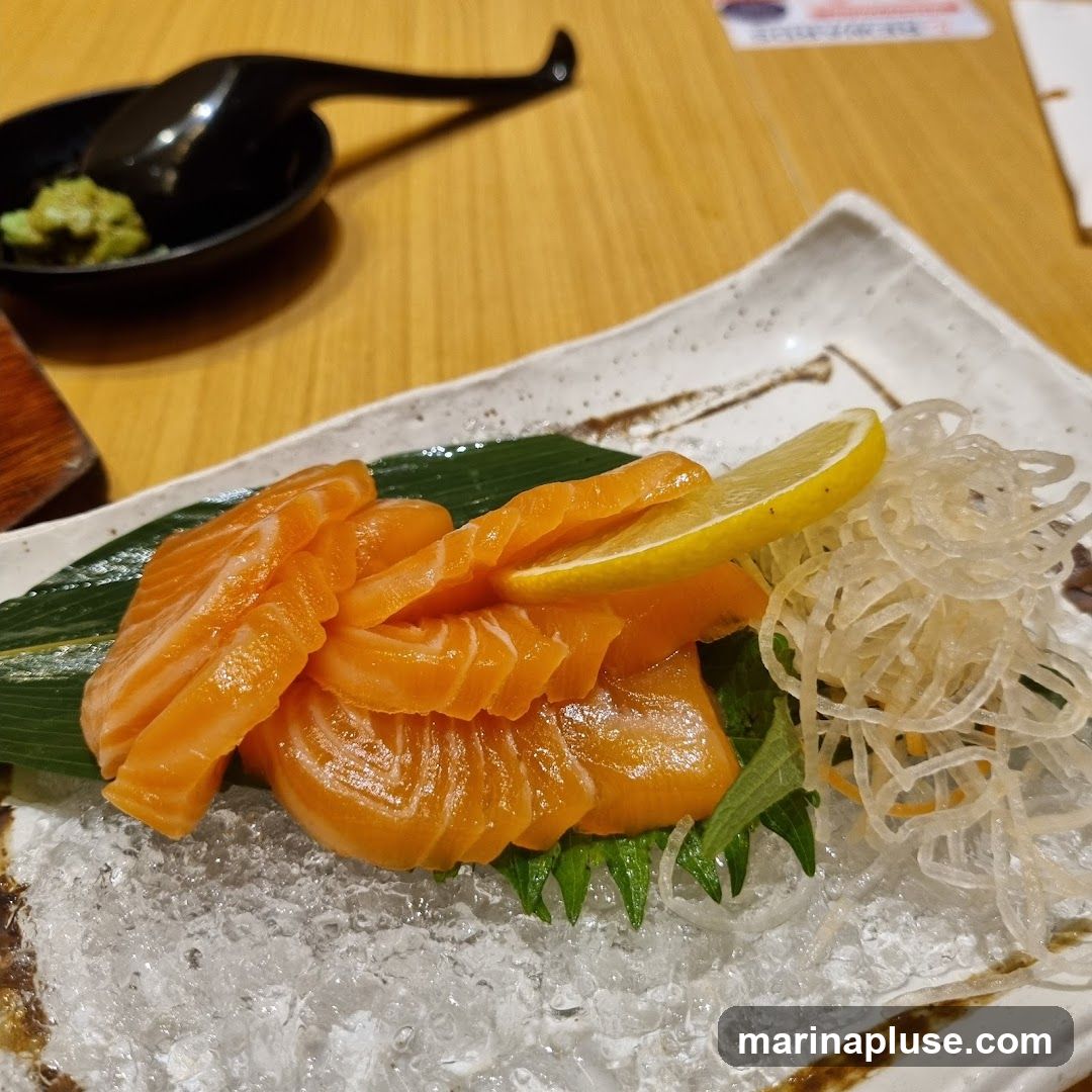 A plate of salmon sashimi served on ice with a lemon slice and shredded daikon radish. A small dish of wasabi and a black spoon are visible in the background.