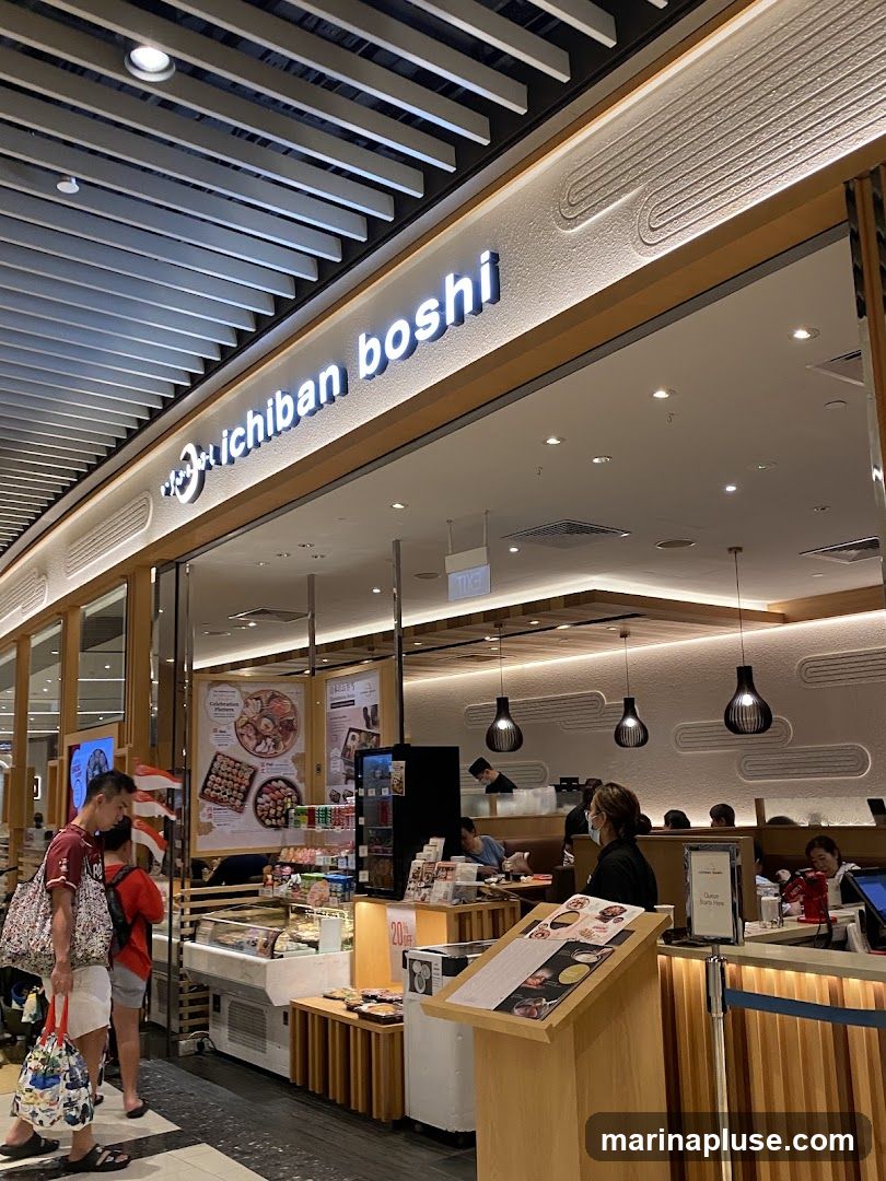 The exterior of the Ichiban Boshi restaurant, showing its illuminated sign above the entrance. Inside, customers are seated at tables, and staff members are visible behind the counter. A display case showcases food items, and promotional materials are placed near the entrance.