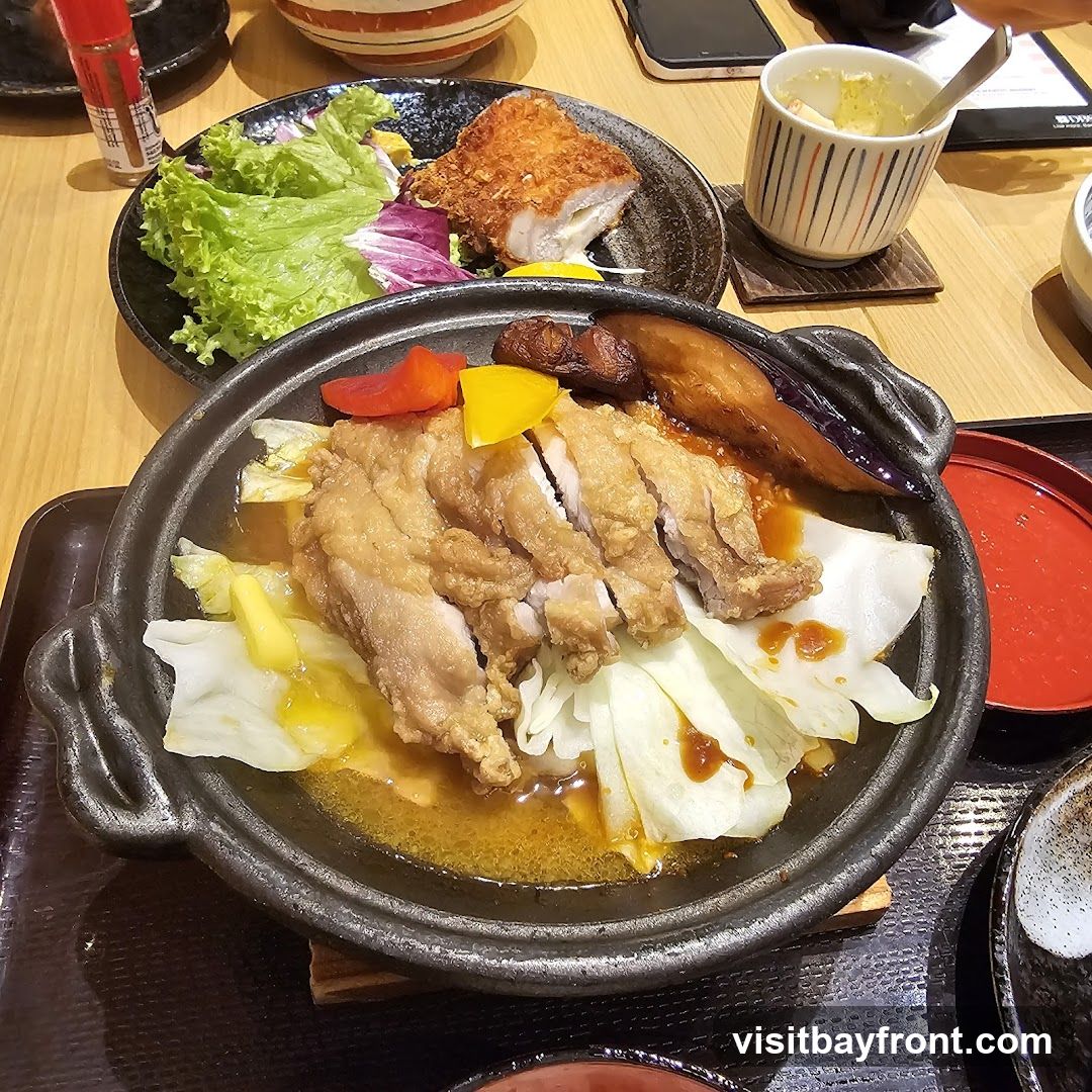 A close-up view of a sizzling dish featuring sliced fried chicken served over cabbage with a savory sauce. Accompanying the chicken are pieces of grilled eggplant and colorful bell peppers. In the background, a plate of fried fish with a side salad and a small cup of what appears to be mashed potatoes or a similar side are visible. The meal is presented on a wooden table.