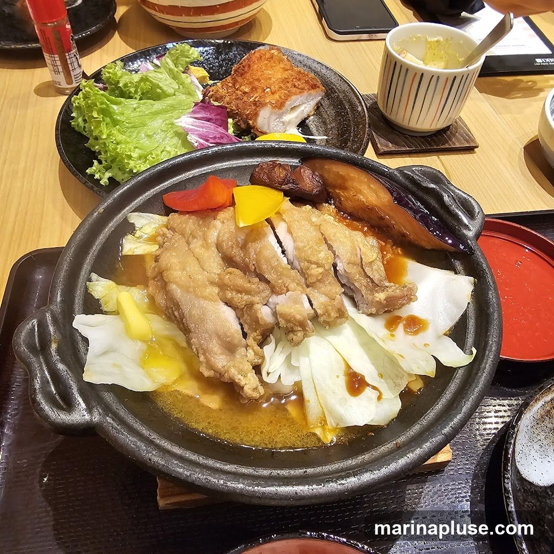 A close-up view of a sizzling dish featuring sliced fried chicken served over cabbage with a savory sauce. Accompanying the chicken are pieces of grilled eggplant and colorful bell peppers. In the background, a plate of fried fish with a side salad and a small cup of what appears to be mashed potatoes or a similar side are visible. The meal is presented on a wooden table.