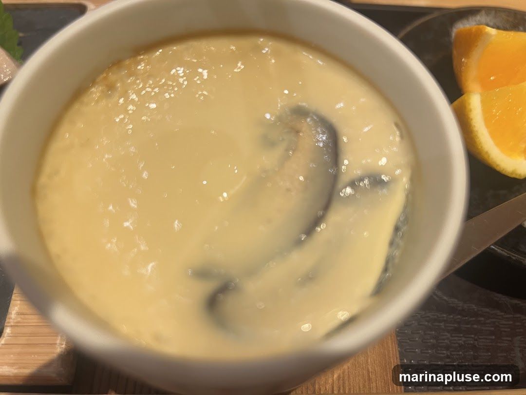 A close-up shot of a white bowl filled with a creamy, light yellow custard. Dark, curved shapes are visible submerged within the custard. To the right of the bowl, two bright orange slices are placed on a dark plate. A utensil, possibly a knife, is also visible on the plate. The overall presentation suggests a dessert or appetizer.