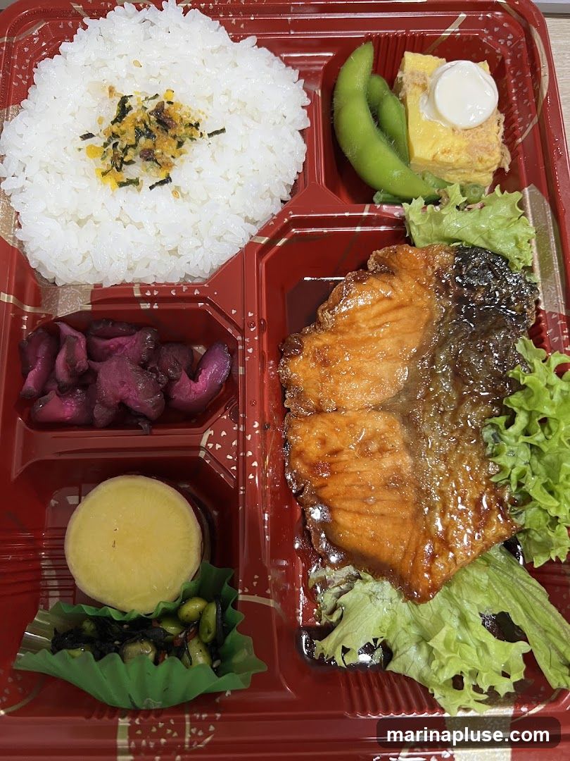 A bento box with rice, grilled salmon, tamagoyaki (rolled omelet), edamame, pickled radish, and a side of seasoned vegetables.