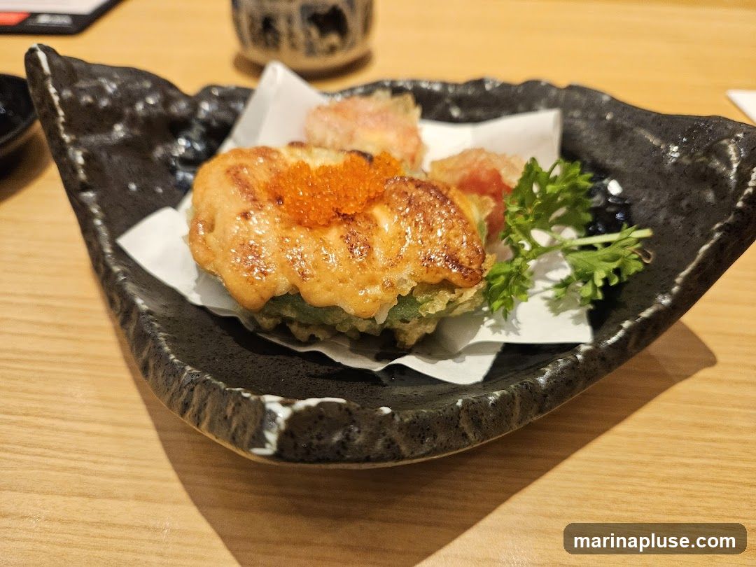 A close-up shot of a tempura dish served on a black, textured plate. The dish features a piece of tempura topped with a creamy sauce and bright orange roe, garnished with a sprig of parsley. The plate rests on a light-colored wooden table.