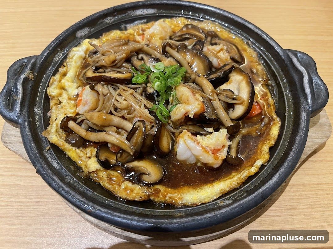 A close-up, overhead view of a sizzling dish featuring a fluffy omelet topped with a generous portion of mixed mushrooms, including shiitake and enoki, and succulent shrimp. The dish is coated in a rich, dark sauce and garnished with chopped green onions. It is served in a black, cast-iron-like dish with handles, resting on a wooden placemat on a wooden table.
