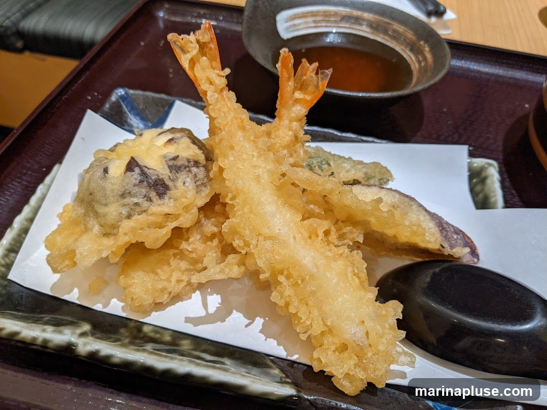 A close-up shot of a tempura platter featuring two large shrimp, a piece of mushroom tempura, and slices of eggplant tempura. A small dish of dipping sauce is visible in the background.