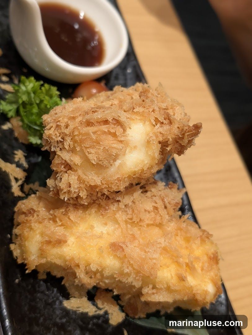 Two pieces of golden-brown fried cheese, coated in crispy panko breadcrumbs, are served on a black plate with a small dish of dark dipping sauce, a sprig of parsley, and a cherry tomato.
