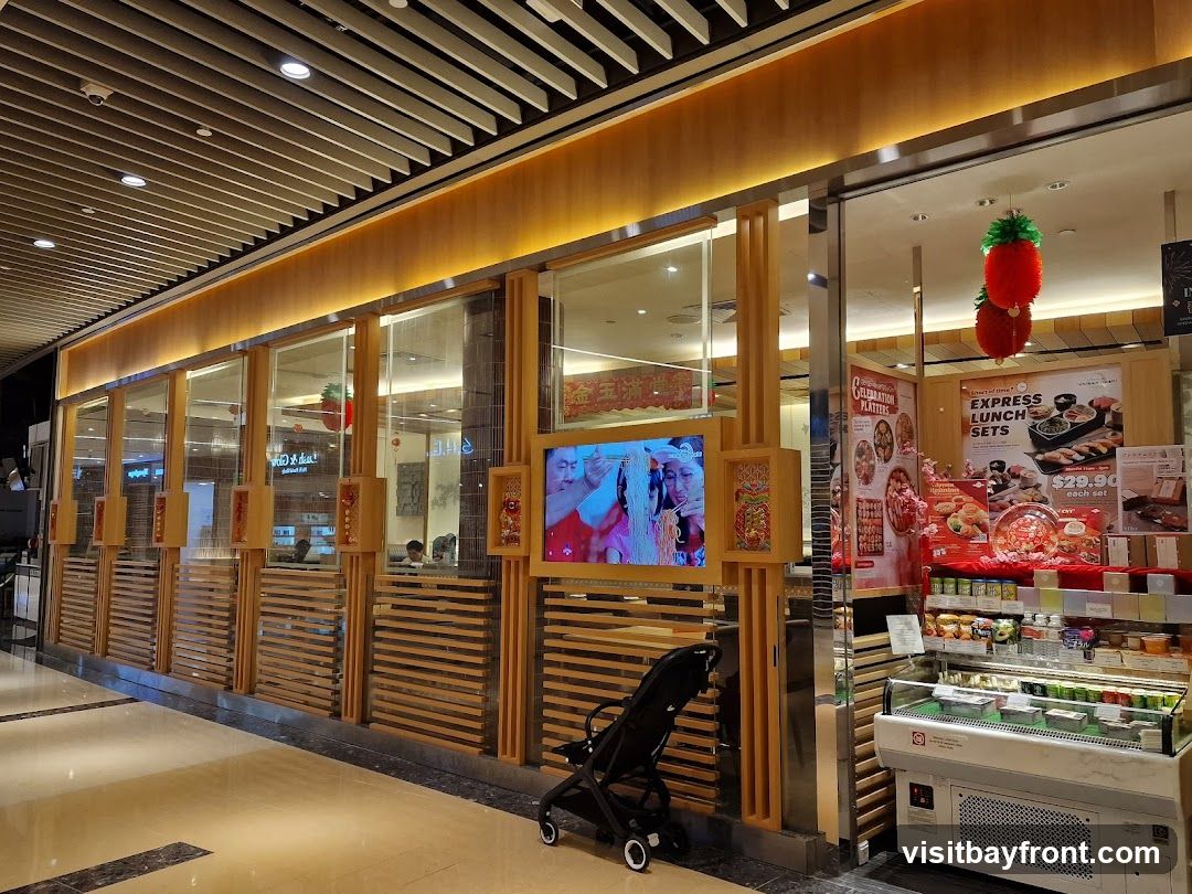 The image shows the interior of a restaurant with a modern, wooden-accented design. A large display case filled with various food items is visible on the right. Promotional posters for "Express Lunch Sets" and "Celebration Platters" are displayed on the wall. A television screen shows images of people. A baby stroller is parked in front of the display. Red, pineapple-shaped decorations hang from the ceiling.