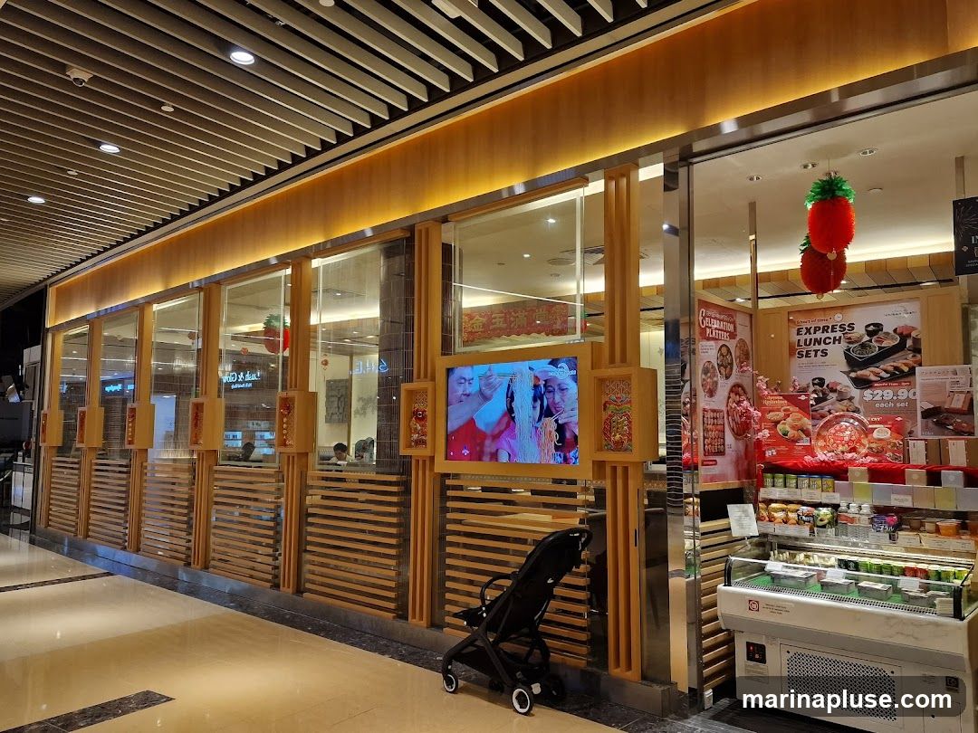 The image shows the interior of a restaurant with a modern, wooden-accented design. A large display case filled with various food items is visible on the right. Promotional posters for "Express Lunch Sets" and "Celebration Platters" are displayed on the wall. A television screen shows images of people. A baby stroller is parked in front of the display. Red, pineapple-shaped decorations hang from the ceiling.