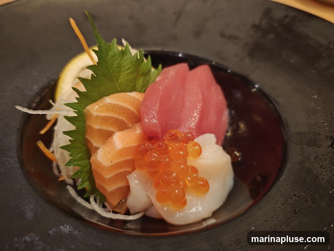 A close-up shot of a sashimi platter featuring slices of salmon, tuna, scallops, and salmon roe, garnished with a lemon slice, shiso leaf, and shredded daikon radish. The food is served in a dark, textured bowl.