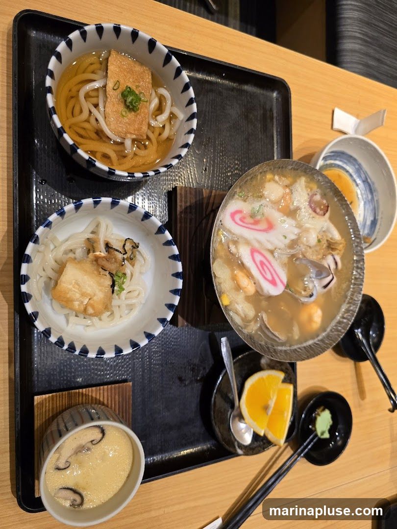 A top-down view of a Japanese food platter on a black tray. The platter includes several bowls of noodles, a bowl of steamed egg custard with mushrooms, and a bowl of seafood soup with fish cakes and shrimp. There are also slices of orange, a small dish of wasabi, and chopsticks.