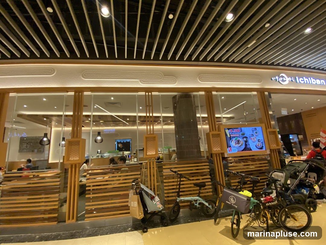 The exterior of a Japanese restaurant with a modern design. The storefront features large glass windows and wooden slatted panels. The restaurant's name, "Ichiban," is visible in Japanese characters and English script above the entrance. Inside, diners can be seen seated at tables. A digital display screen shows food items and prices. Several bicycles and a stroller are parked outside the entrance.