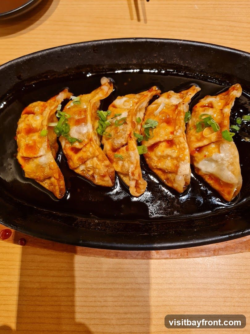 A close-up shot of pan-fried dumplings served in a black oval dish with a dark sauce and garnished with chopped green onions. The dumplings are golden brown and appear crispy on the bottom. The dish is placed on a wooden table.