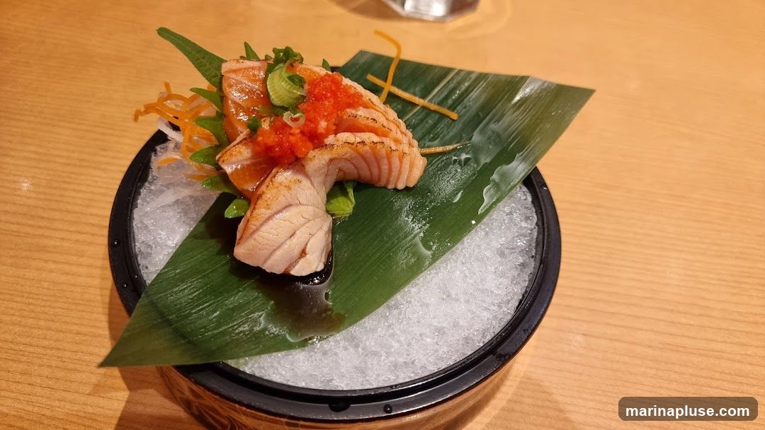 A close-up shot of salmon sashimi served on a bed of crushed ice, garnished with a green leaf, shredded carrots, and a dollop of red roe and chopped scallions. The dish is presented in a black bowl on a wooden table.