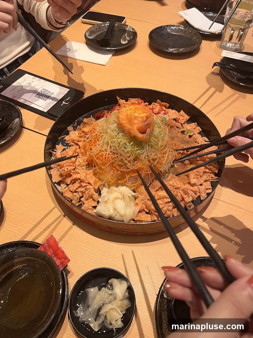 A group of people are about to toss a Yee Sang platter, a traditional Chinese New Year dish. The platter is filled with shredded vegetables, crispy crackers, and topped with salmon. Chopsticks are poised over the dish, ready to mix the ingredients. Small bowls of pickled ginger and dipping sauce are also visible on the wooden table. A menu or order form is placed on the table, showing a QR code.