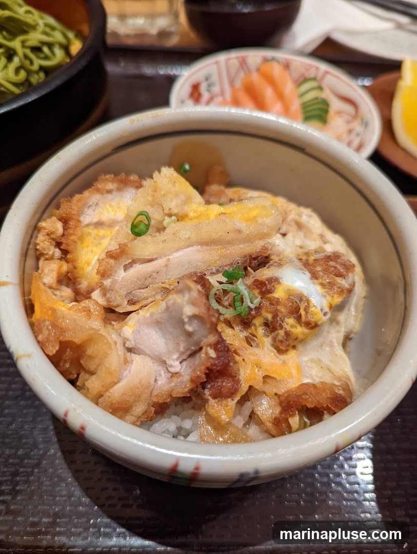 A close-up shot of a chicken katsu donburi, a Japanese rice bowl dish. The bowl is filled with white rice topped with pieces of crispy fried chicken cutlet, simmered with egg and onions. Garnished with chopped green onions. In the background, there are other dishes including green noodles and sashimi.