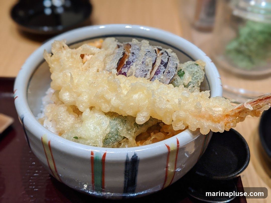 A close-up shot of a tempura rice bowl. The bowl contains white rice topped with various tempura pieces, including a large shrimp, eggplant, and broccoli. The bowl has a decorative rim with vertical stripes in red, green, and black. A small dark dish is visible to the right of the bowl. The background is blurred, showing a wooden table and some out-of-focus elements.