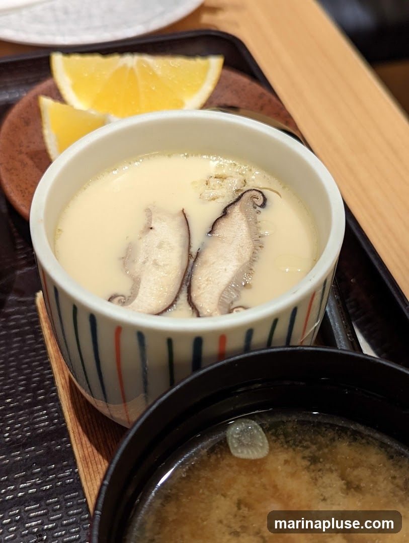 A close-up shot of a Japanese meal, featuring a bowl of chawanmushi (savory steamed egg custard) topped with shiitake mushrooms and garnished with two lemon wedges. A bowl of miso soup is visible in the foreground.