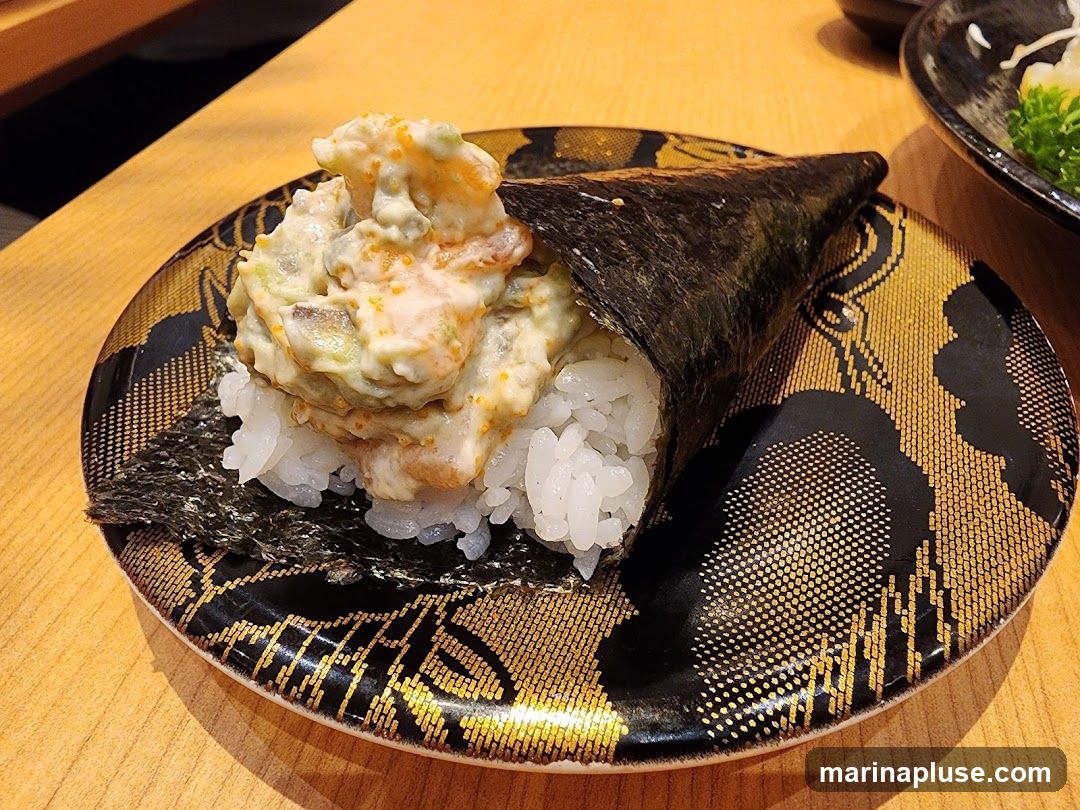 A close-up shot of a spicy tuna hand roll on a decorative black and gold plate. The hand roll is made with sushi rice, nori seaweed, and a creamy, spicy tuna filling mixed with avocado and tobiko. The plate has an intricate pattern of gold lines and dots.