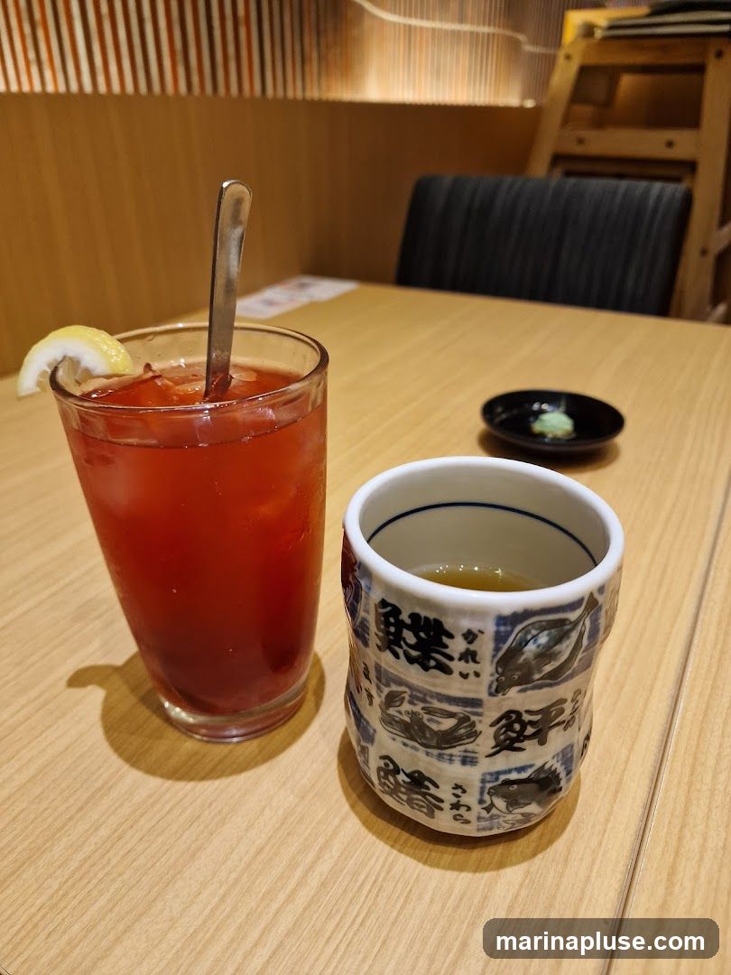 A tall glass of red iced beverage with a lemon slice and a spoon, and a small cup of tea with Japanese-style illustrations of fish and crabs. A small dish with a green condiment, likely wasabi, is also visible on the wooden table. The background shows a restaurant interior with wooden paneling and seating.
