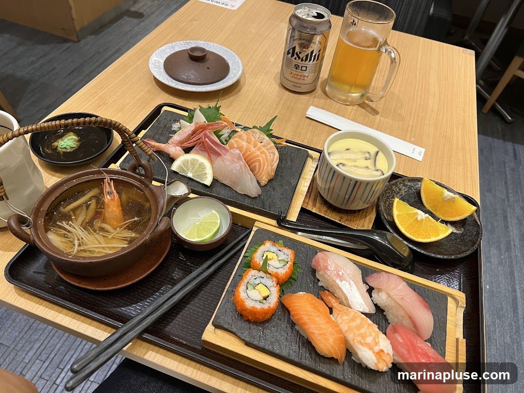 A wooden table set with a Japanese meal. The meal includes a pot of soup with a shrimp and mushrooms, a plate of assorted sashimi (salmon, white fish, shrimp), a bowl of chawanmushi (steamed egg custard) with mushrooms, a plate of sushi rolls and nigiri (salmon, white fish, tuna, shrimp), a can of Asahi beer, and orange slices. Chopsticks and a small dish of wasabi are also visible.