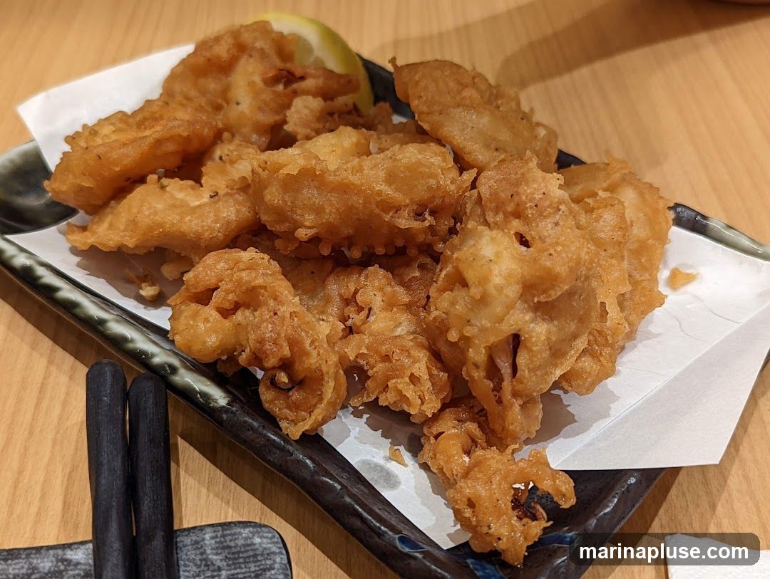 A plate of golden-brown fried calamari served with a lemon wedge and white paper liner. Two dark chopsticks rest on a chopstick holder in the foreground. The calamari appears crispy and is piled high on a dark, textured plate.