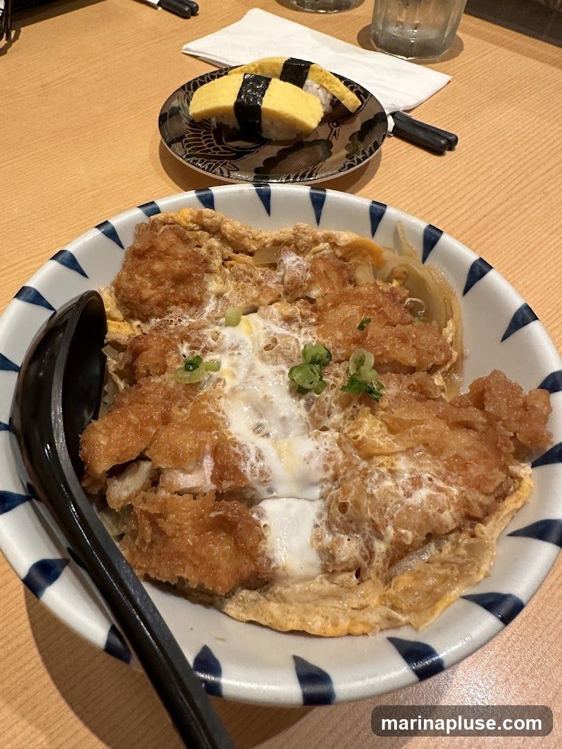 A close-up shot of a bowl of katsudon, a Japanese dish consisting of a deep-fried pork cutlet, egg, and thinly sliced onions simmered in a savory sauce, served over rice. In the background, two pieces of tamagoyaki sushi, a rolled omelet, sit on a decorative plate. The meal is presented on a wooden table with chopsticks and a glass of water.