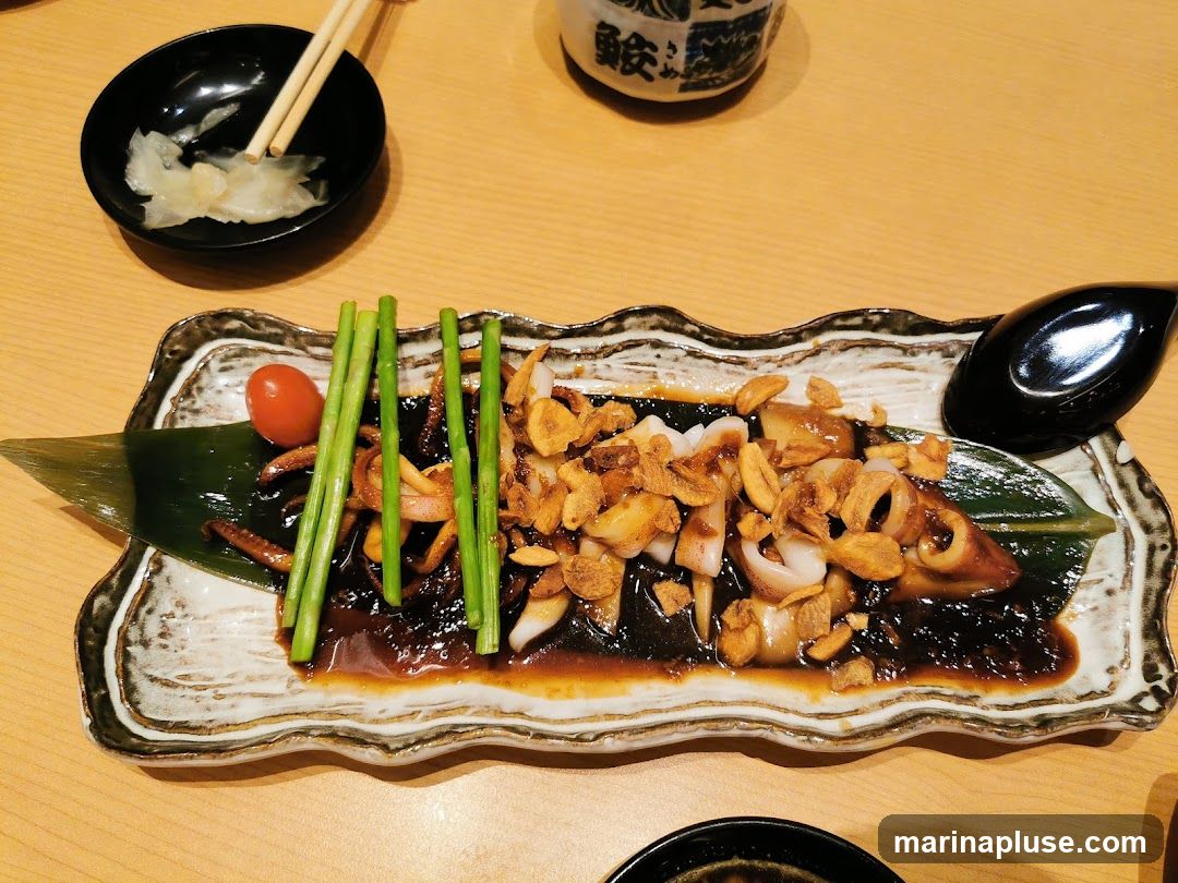 A close-up shot of a Japanese-style dish featuring tender squid rings and tentacles, coated in a dark, savory sauce and topped with crispy fried garlic. The dish is garnished with a single cherry tomato and several stalks of bright green asparagus, all served on a decorative platter with a banana leaf base. A small black bowl with pickled ginger and chopsticks is visible in the upper left corner, and a dark spoon rests on the right side of the platter. The meal is presented on a light wooden table.