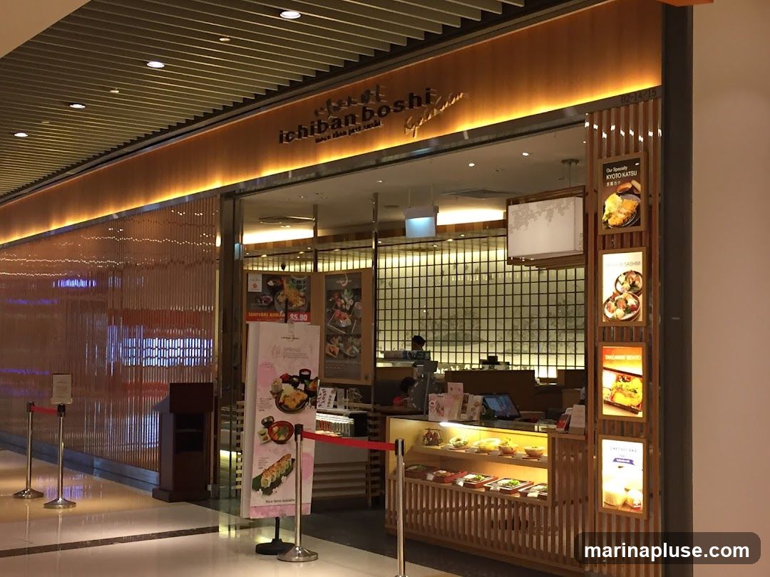 The exterior of a Japanese restaurant called Ichiban Boshi. The restaurant has a warm, inviting ambiance with wooden accents and soft lighting. A large sign with the restaurant's name is visible above the entrance. Several framed pictures of food items are displayed on the right side of the entrance, showcasing dishes like Kyoto Katsu, Sashimi, Bento, and Cheesecake. Inside, a display case shows various food items, and a menu board is visible.